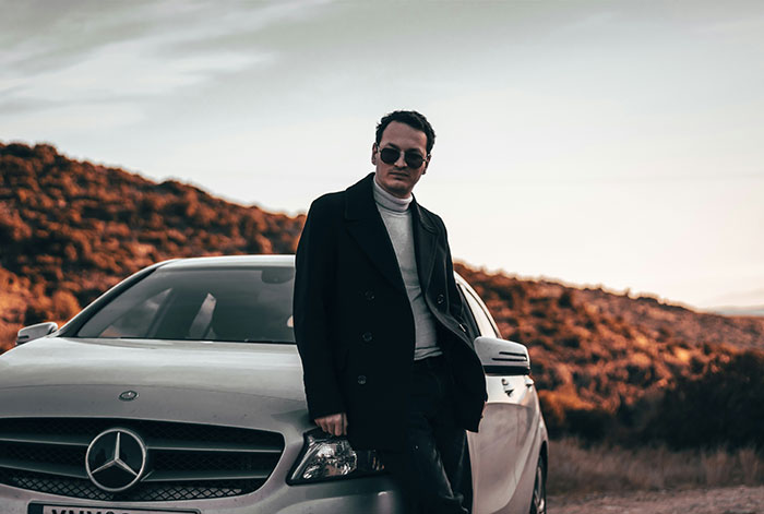 Man in sunglasses leaning against a white Mercedes-Benz in a natural setting, reflecting on popping the question and embarrassment.
