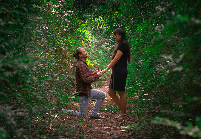 Man popping the question on one knee in a forested path surrounded by greenery during a marriage proposal moment.