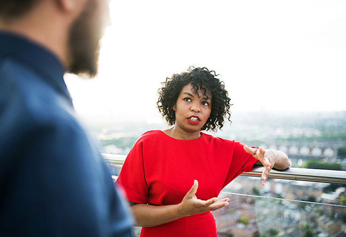 Woman in red explaining nervously to a man on rooftop, capturing engagement proposal embarrassment and awkward moment.