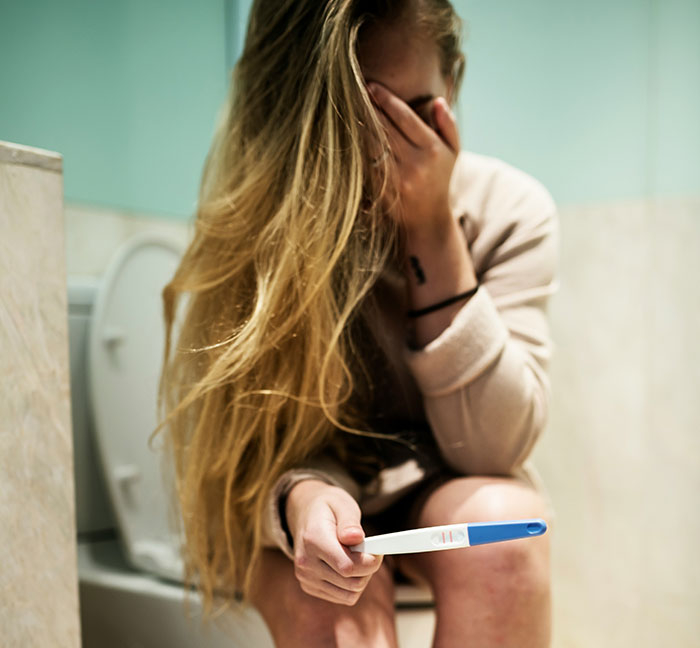 Young woman sitting on a toilet, looking distressed while holding a pregnancy test, capturing an embarrassing moment.