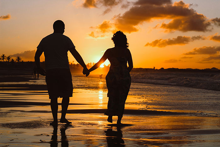 Couple holding hands walking on the beach at sunset, symbolizing moments of popping the question and ring embarrassment.