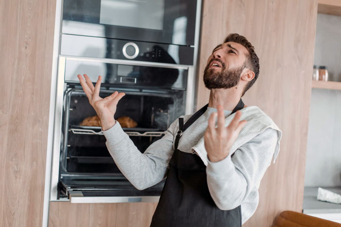 Frustrated man in a kitchen wearing an apron, reacting to burnt steak cooked in the oven with his hands raised.