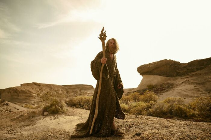 Character in The Rings of Power season 3 wearing a cloak and holding a staff in a desert landscape at sunset.