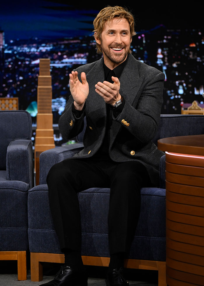 Ryan Gosling smiling and clapping hands during a late night talk show interview in a dark blazer and black pants.