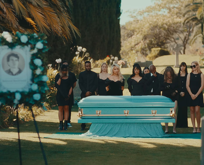 Mourners dressed in black stand around a turquoise coffin in a solemn outdoor scene from Sabrina Carpenter music videos.