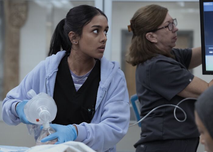 A medical professional wearing gloves works with equipment while another checks a monitor in The Pitt Season 2, Episode 12.