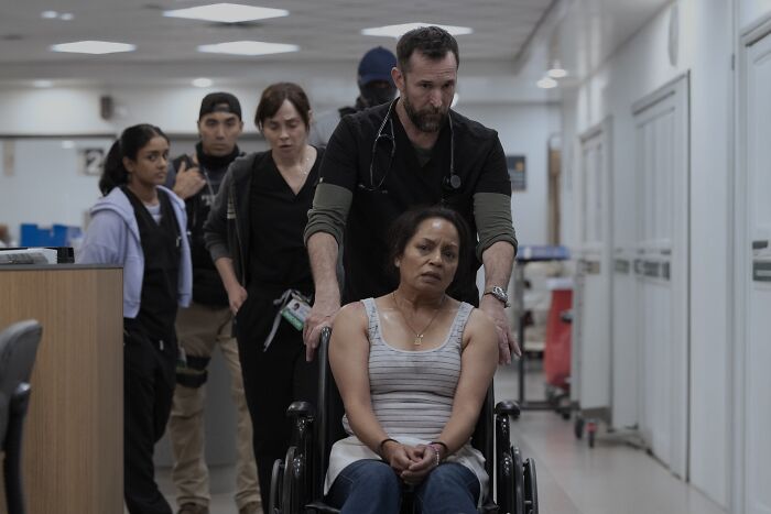 A man pushes a distressed woman in a wheelchair down a hospital hallway with medical staff following in The Pitt season 2.