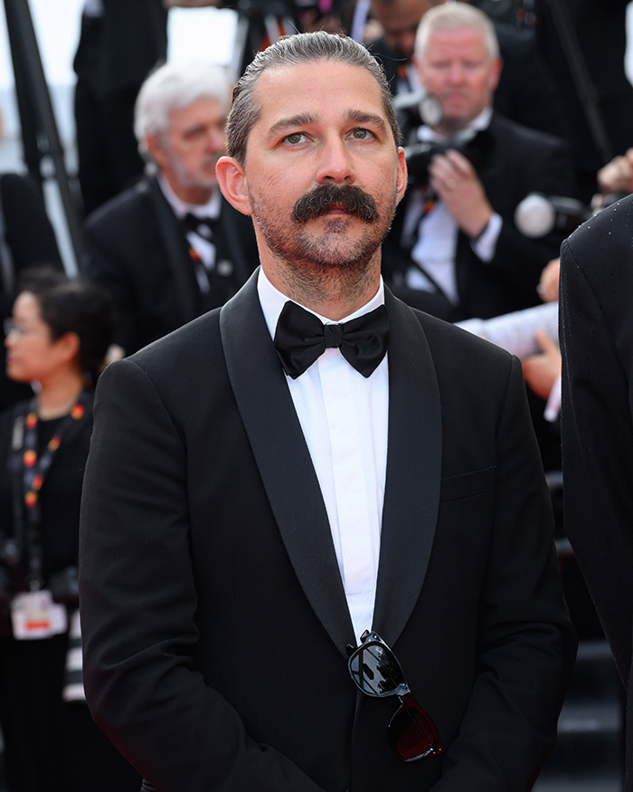 Shia LaBeouf in a black tuxedo with a bow tie at a formal event, looking serious with a mustache and slicked-back hair.