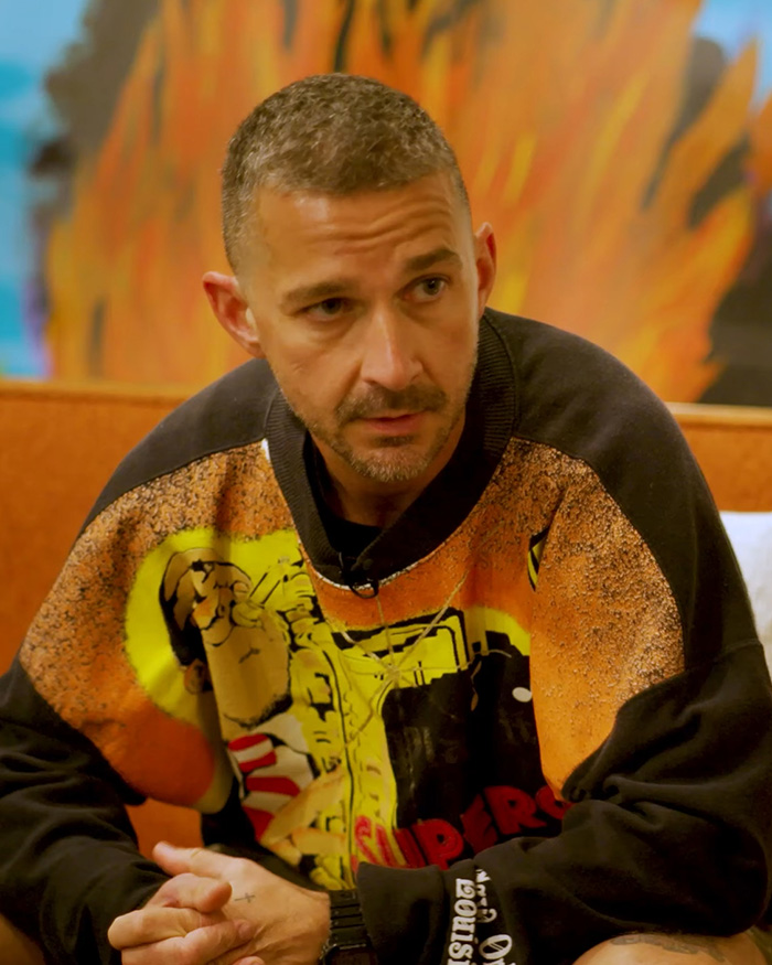 Shia LaBeouf sitting indoors wearing a colorful sweatshirt, looking serious in new footage from Rome incident.