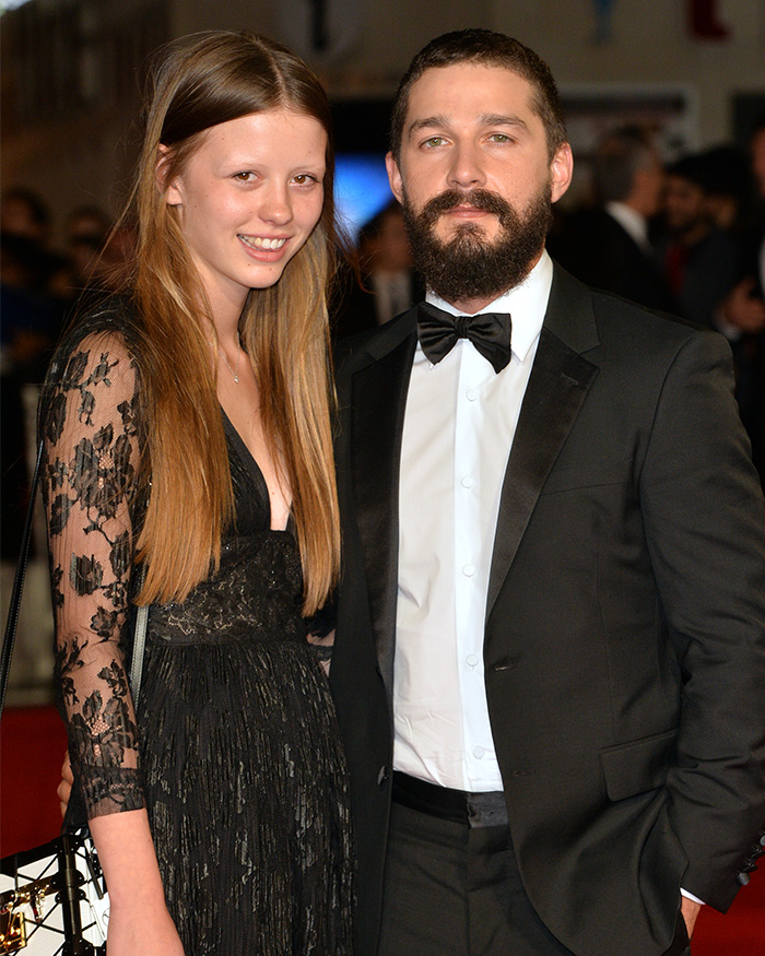 Shia LaBeouf in a black tuxedo with a woman in a black dress at a formal event, posing for photos.