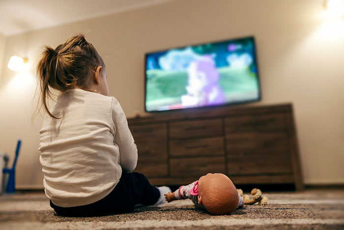 Child watching TV with a doll nearby, evoking childhood memories and moments people often remember from youth.