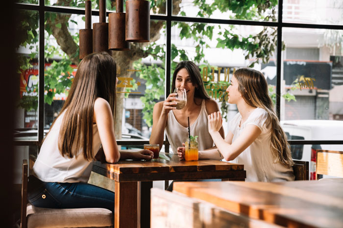 Three young women having drinks and chatting at a cafe, illustrating mom using sis as a free sitter concept.