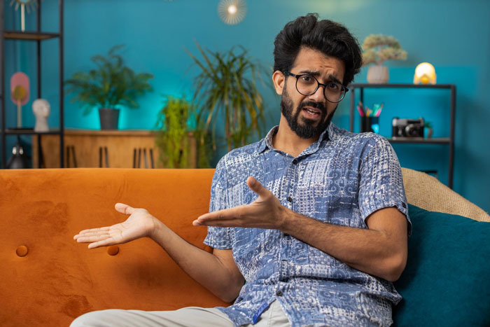 Man with glasses and beard sitting on orange couch, expressing shock and confusion about woman giving baby her last name.