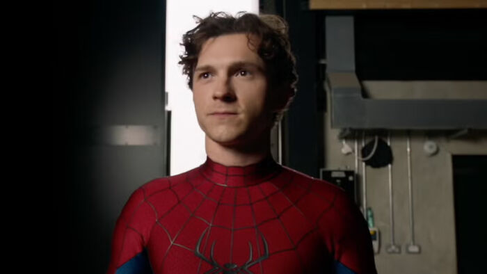 Tom Holland as Spider-Man in his red and blue suit, facing forward with a serious expression in a dimly lit room.
