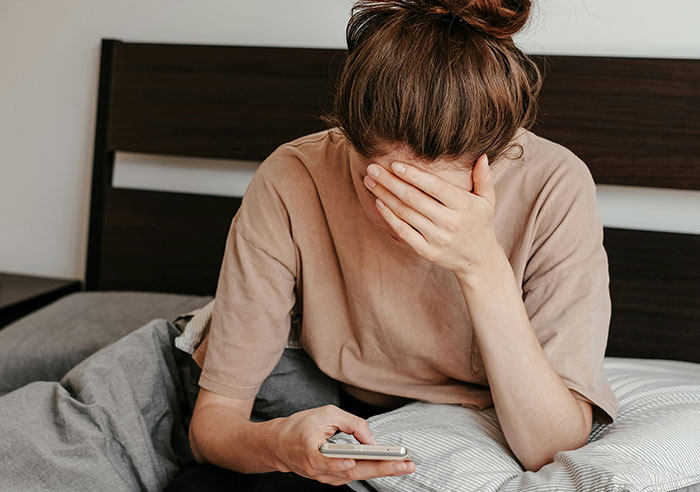Woman sitting on bed, covering face with hand while looking at phone, reflecting on moments that made her feel spoiled