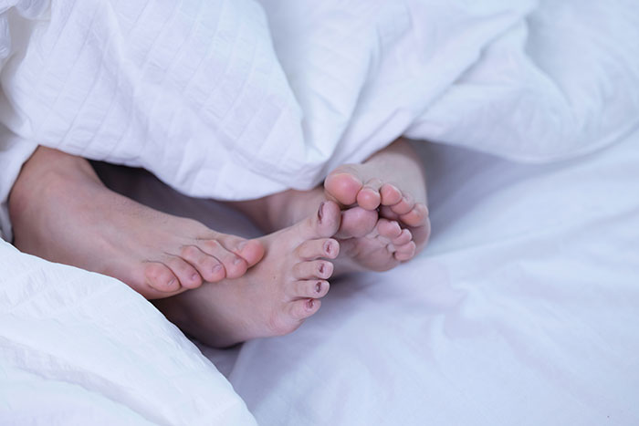 Two pairs of bare feet tangled together under white bed sheets, illustrating a relationship scene.