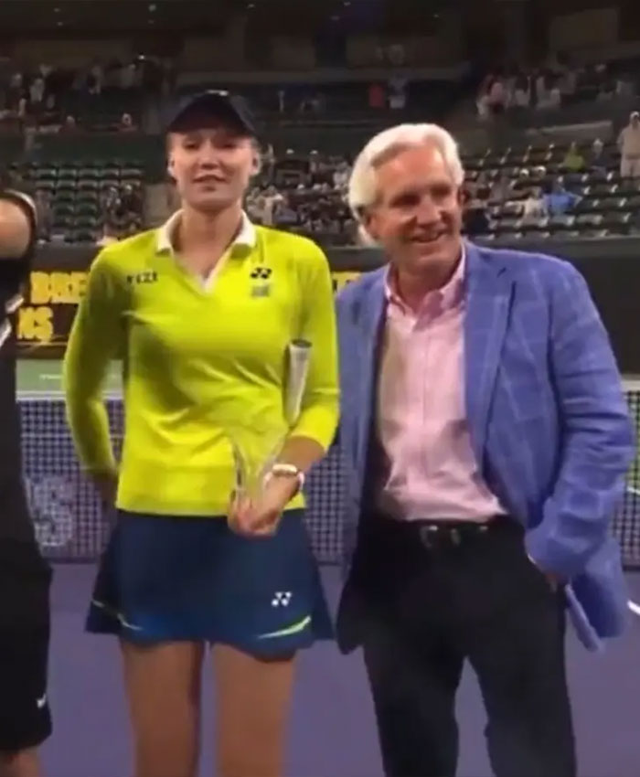 Elena Rybakina holding trophy on tennis court while awkwardly pushing presenter’s hand away during ceremony