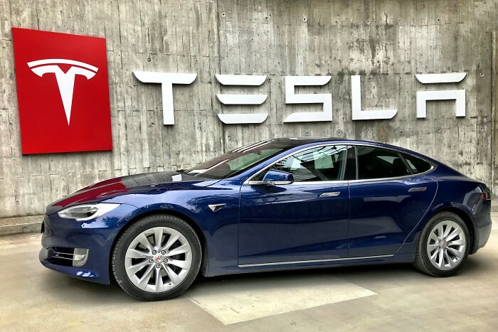 Blue Tesla electric car parked in front of concrete wall with Tesla logo, representing shaky industries at risk of collapse.