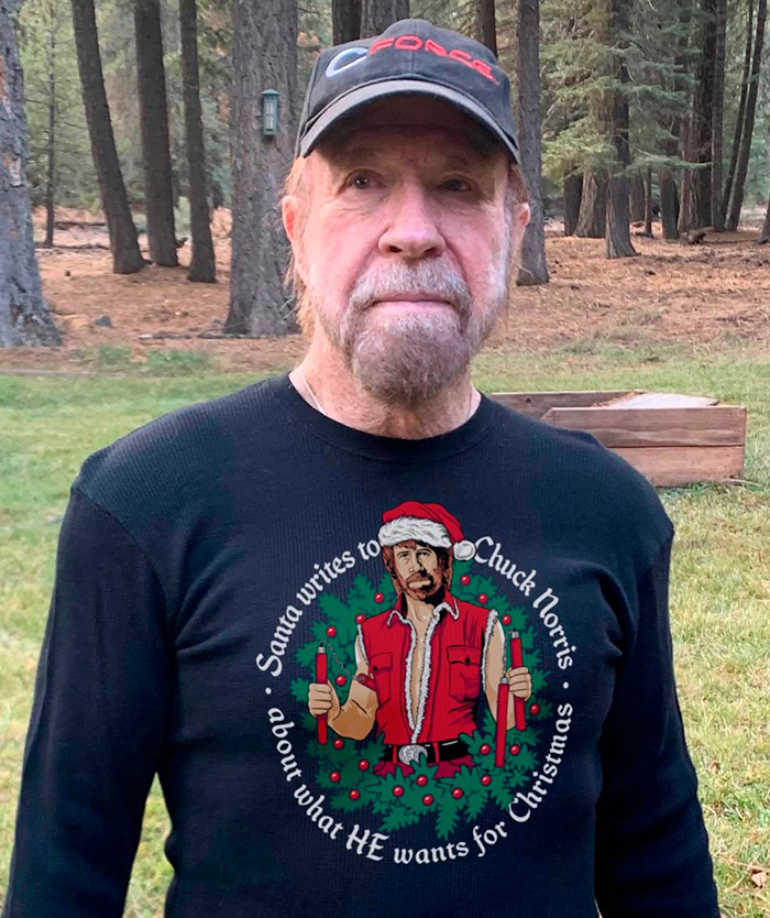 Man wearing a black shirt with a humorous Chuck Norris Christmas design standing outdoors among trees.