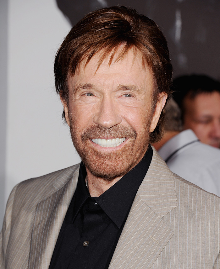 Chuck Norris smiling at an event, wearing a tan suit jacket and black shirt, inspiring popular memes tribute.