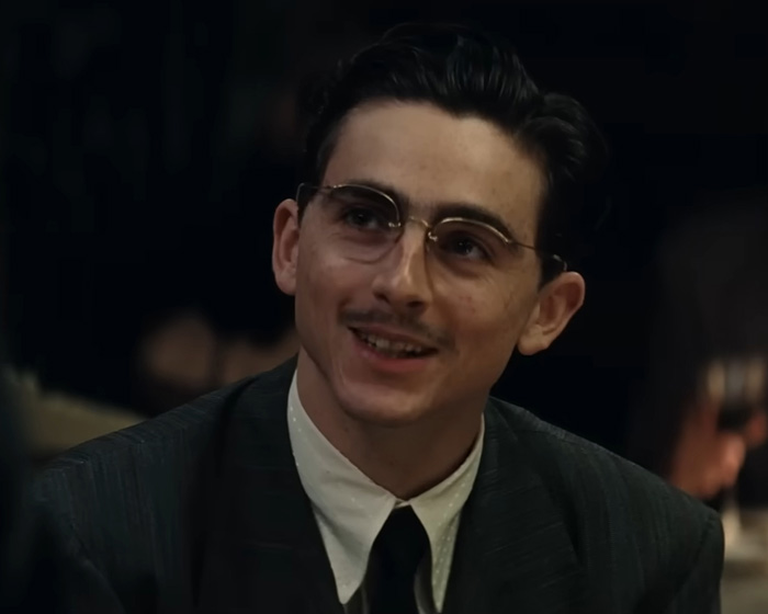 Timoth&eacute;e Chalamet wearing glasses and a suit, smiling during a conversation amid opera and ballet controversy.