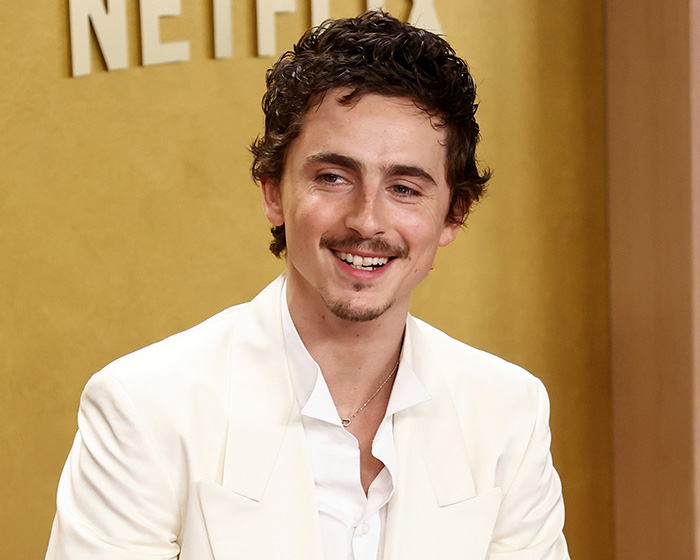 Timoth&eacute;e Chalamet smiling in a white suit during an interview amid backlash over opera and ballet comment.