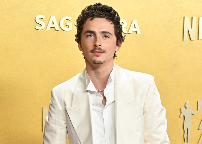 Timoth&eacute;e Chalamet in a white suit posing at the SAG Awards event against a gold background with SAG logo.