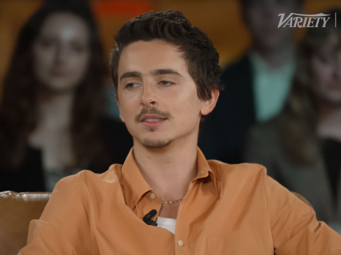 Timoth&eacute;e Chalamet in a seated interview, wearing an orange shirt, amidst a discussion about ballet and opera remarks.