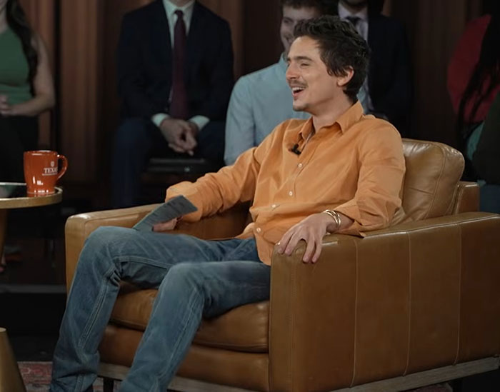 Timoth&eacute;e Chalamet in a casual interview setting, wearing an orange shirt and jeans, discussing ballet drama.