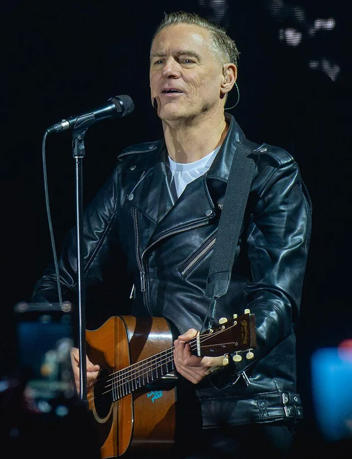 Man in a black leather jacket playing guitar and singing on stage, illustrating humans neurologically entering adulthood at 32.