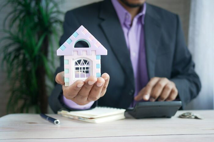 Man in a suit holding a model house with a calculator on the table, symbolizing shaky industries and economic risks.