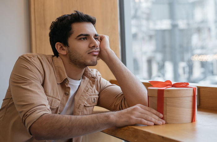 Young man looking disappointed while sitting by a window with a wrapped gift, reflecting on trashing girlfriend's gift.