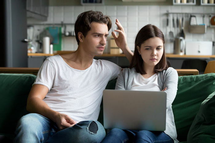 Young couple on a couch with a laptop, showing tension and frustration related to trashing girlfriend's gift.