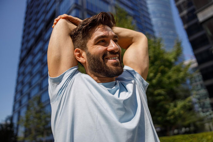 Man in a light blue shirt stretching outdoors, smiling with skyscrapers and greenery in the background, trashing girlfriend's gift theme.