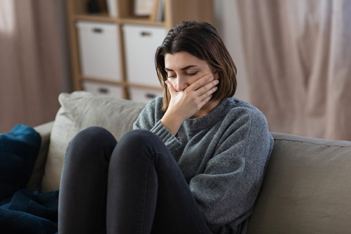 Young woman sitting on couch covering mouth, looking upset and reflecting on trashing girlfriends gift at home.
