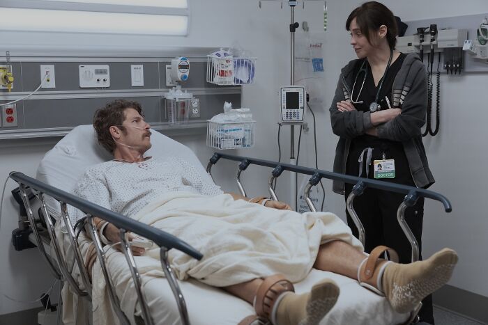 Hospital scene from The Pitt season 2 episode 13 with patient in bed and doctor standing nearby in conversation.