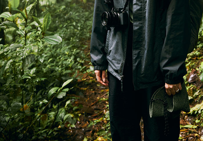 Person wearing a dark jacket with a camera around their neck, holding a rolled map in a dense green forest setting.