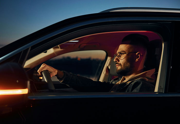 Man wearing glasses driving a car at dusk, focused and calm, illustrating one-in-a-million stories that people lived through.