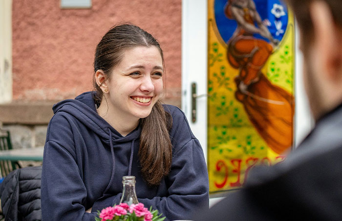 Young woman smiling and sharing one-in-a-million stories in an outdoor cafe setting with a colorful mural behind her.