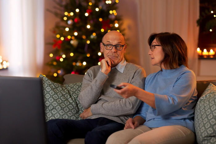 Couple sitting on couch sharing unconventional family traditions during holiday season with decorated Christmas tree behind them.
