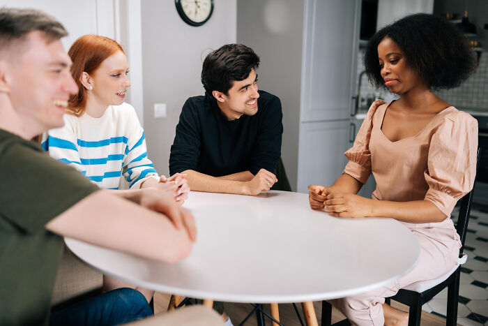 Four friends sharing stories about unconventional family traditions while sitting around a white table indoors.