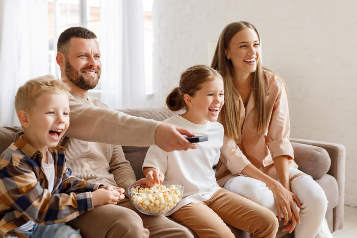 Family enjoying unconventional family traditions together on a couch, sharing popcorn and watching TV happily.