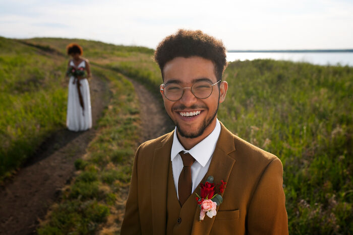 Young man smiling outdoors in a brown suit with a boutonniere, capturing unconventional family traditions in a natural setting.