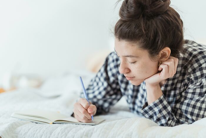 Young woman in a checkered shirt lies on bed, writing in notebook, reflecting on challenging beliefs and controversial opinions.