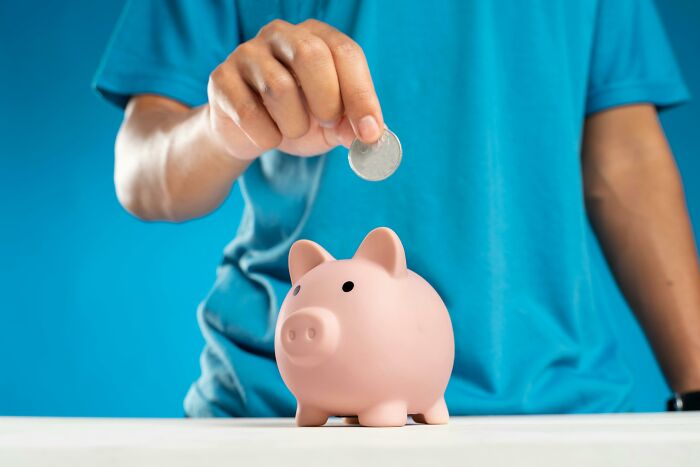 Person in blue shirt holding coin above piggy bank, symbolizing challenge your beliefs on saving and spending opinions.