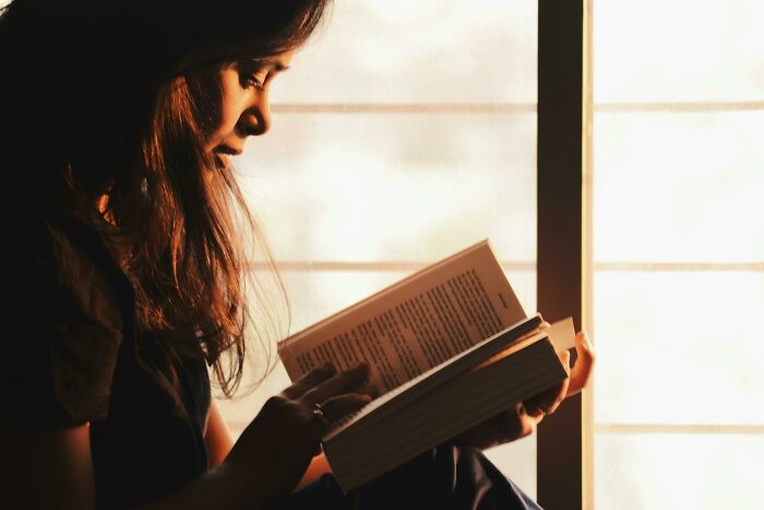 Young woman reading a book by the window, reflecting on challenging beliefs and controversial opinions.