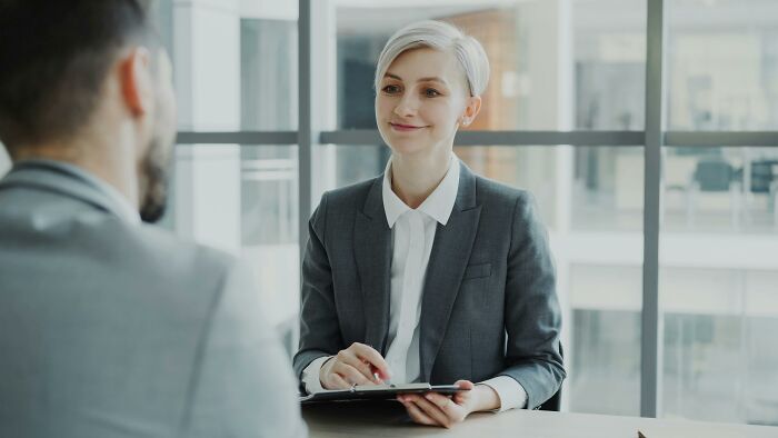 Professional woman in a gray suit holding a tablet, engaged in a discussion challenging beliefs in an office setting.