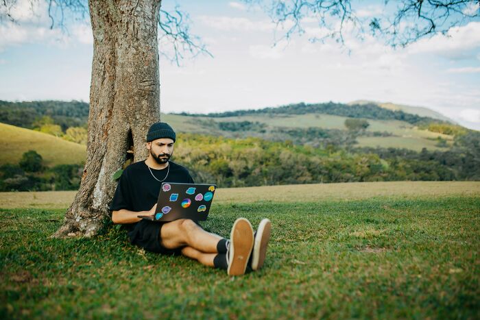 Man sitting under a tree using a laptop in a field, reflecting and challenging beliefs on controversial opinions.