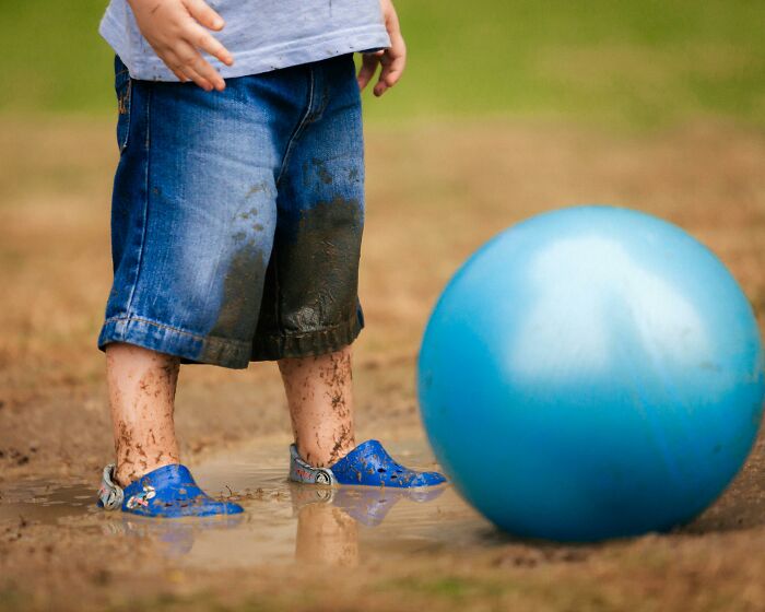 Child standing in muddy water with dirty shorts and blue shoes near a large blue ball, challenging beliefs concept.