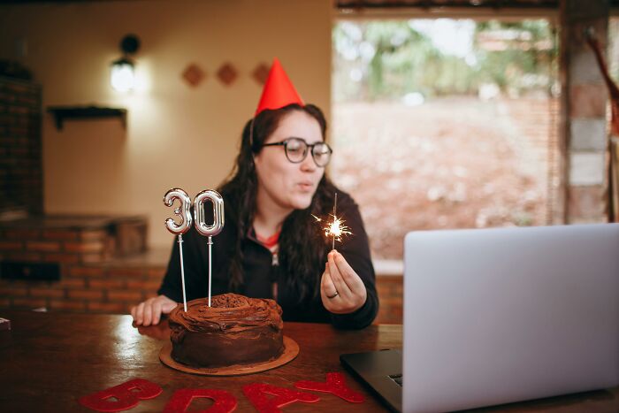 Woman in a red party hat celebrating birthday with chocolate cake and sparklers during a virtual online party session.